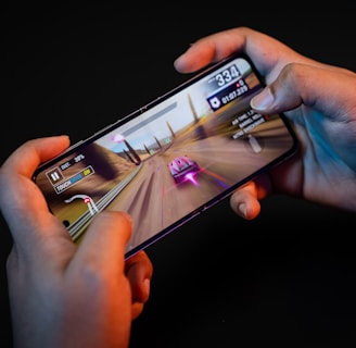 Hands holding a smartphone with a racing game displayed on the screen. The image shows a vibrant racing scene with a red and white car moving at high speed along a desert-like track. Various on-screen controls and speed indicators are visible.